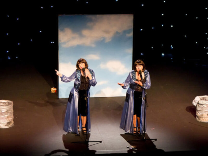 Actrices interpretando en escenario con fondo de cielo y luna, vestidas con capas azules y negras.