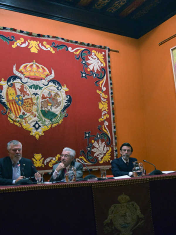 Tres hombres sentados en una mesa con micrófonos, en un salón con tapiz y cuadro decorativo en la pared.