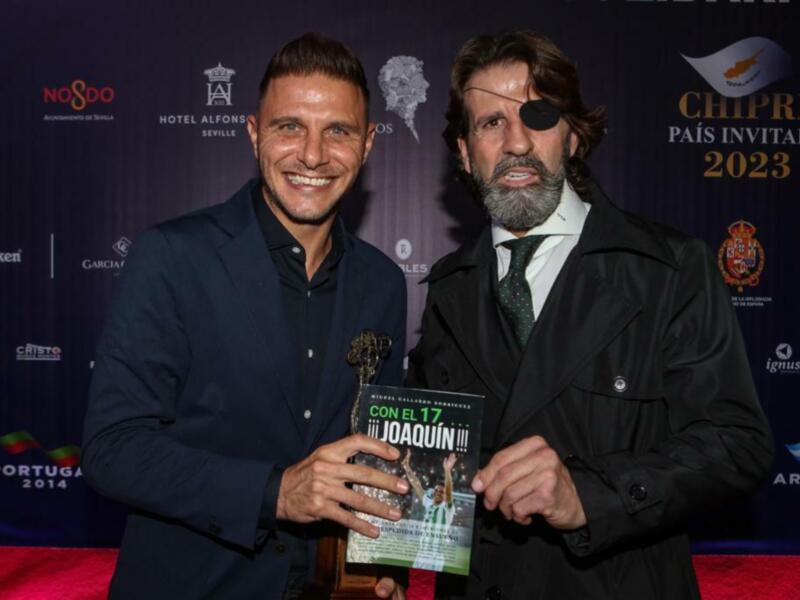Dos hombres posan en una alfombra roja con un fondo de patrocinadores, uno sostiene un libro titulado '¡¡¡JOAQUÍN!!!'.