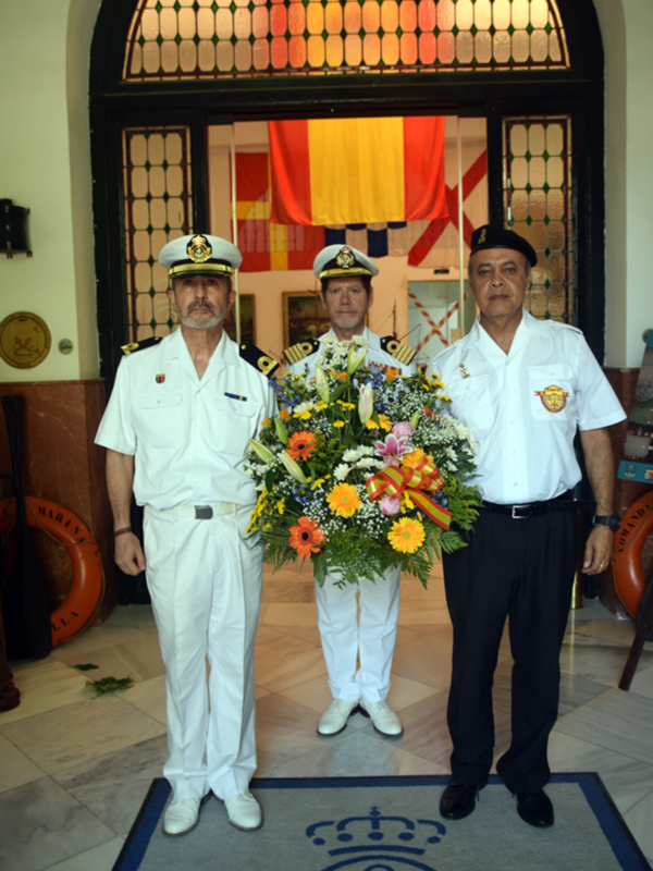 Tres hombres de uniforme militar de la Armada de España, con uniforme blanco y un gran bouquet de flores en la mano de uno de ellos, en el interior de un edificio con un arco de vidrio y un tapiz de la bandera de España colgando en la parte de a...